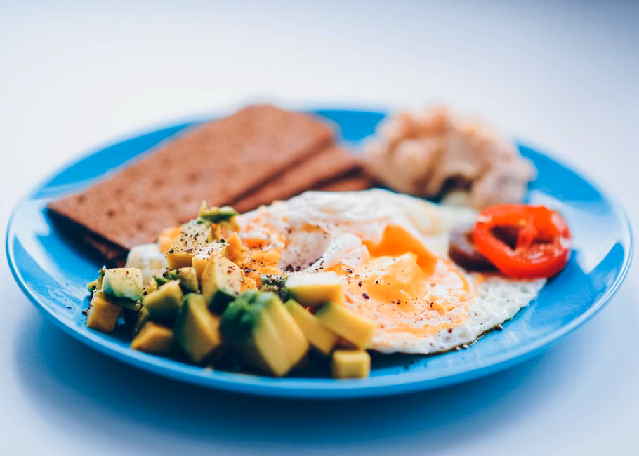 Bright and colorful breakfast featuring fried eggs, avocado, and fresh vegetables on a blue plate.